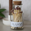 Glass jar with cork lid containing matches labeled 'Love at first light' on a neutral background.