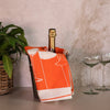 Bottle of champagne in a wine bucket with an orange and cotton towel with a plant and glasses in the background.