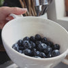 Off white ceramic berry colander being used to rinse blueberries under unning water